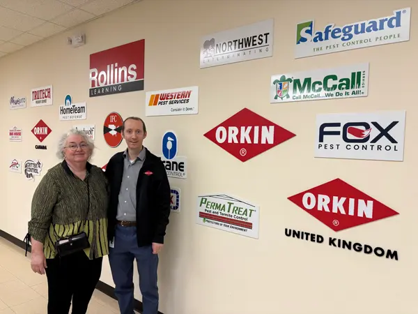 Orkin entomologists and mother and son Patricia and Ben Hottel standing together beside a wall featuring Rollins pest control brand logos such as Orkin, Safeguard, Northwest Exterminating, McCall Service, Fox, Western Pest Services, and PermaTreat.