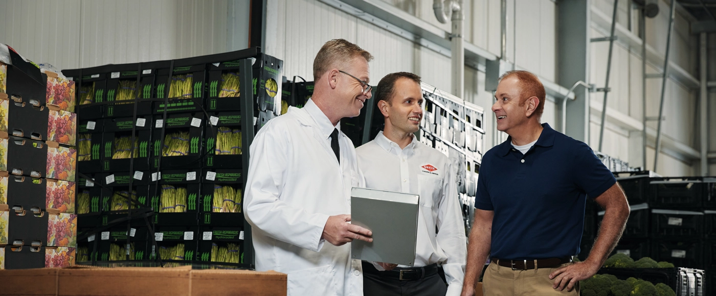 An Orkin Pro with two professional men, including one in a white lab coat holding a tablet, standing and conversing in a large warehouse filled with stacked crates.