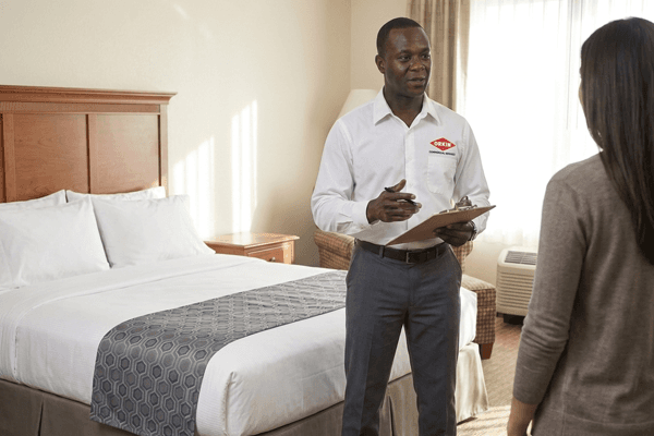 A Commercial Orkin Pro holding a clipboard speaks with a client inside a hotel room near a made bed, discussing bed bug service.