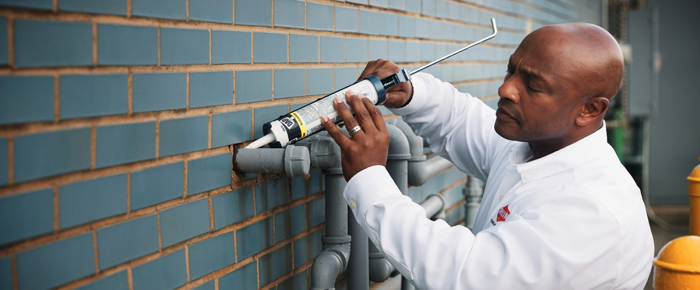 Close-up of an Orkin Pro in a white uniform shirt using a caulking gun to seal gaps around grey utility pipes on an exterior blue brick wall.